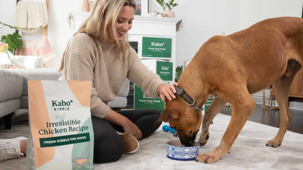 Kabo showing their fresh kibble with a woman petting a dog in the background.